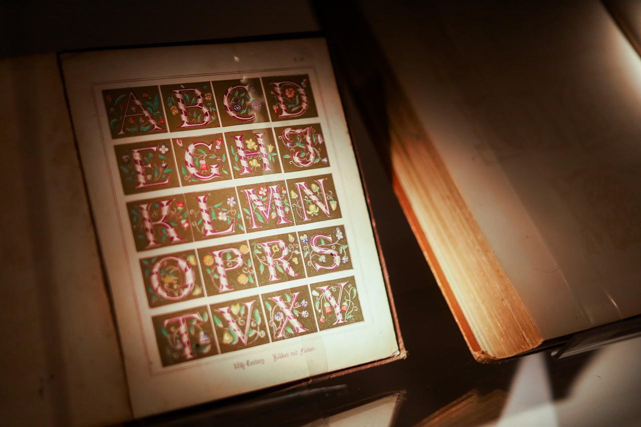An open book, displaying an elaborate illustration of the alphabet. 