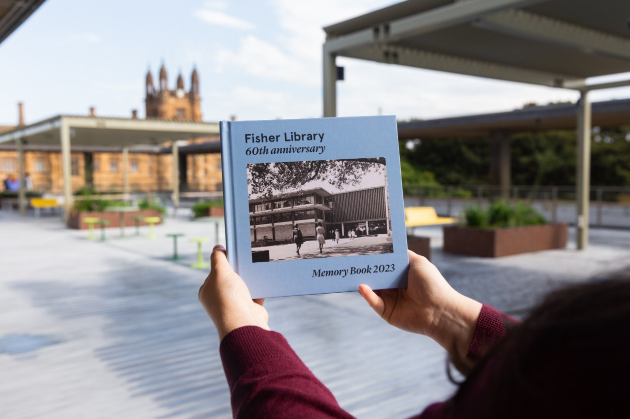 Person holding Fisher Library 60th anniversary book with the Fisher Library roof terrace in the background