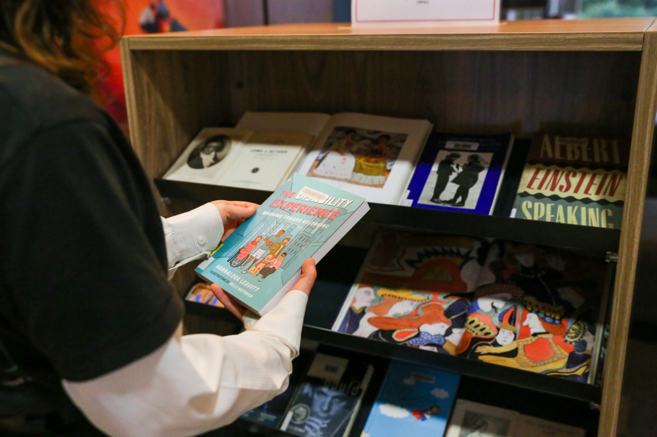 Person browsing a bookshelf, holding a book entitled 'The Disability Experience'