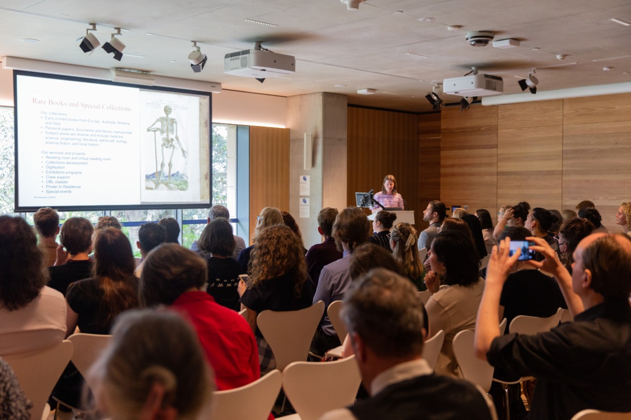 An audience of people looking at a single presenter and screen projection