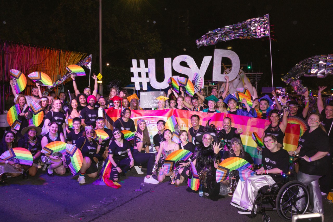 Large group of people smiling and holding pride progress flags. Colourful and sparkly streamers in the background.