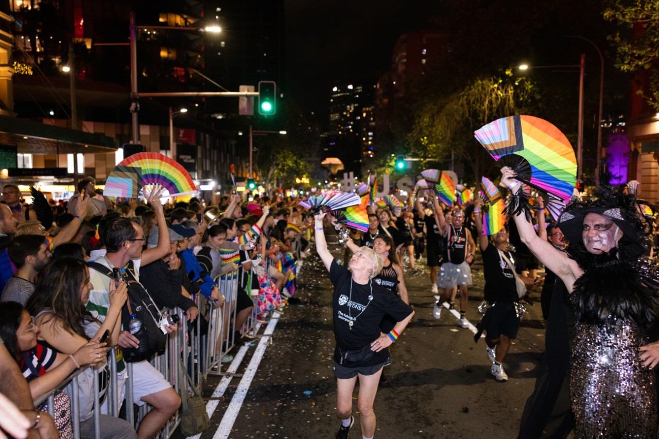  Mardi Gras marchers walk down Oxford st waving pride progress flags. Busy crowds watch on. 