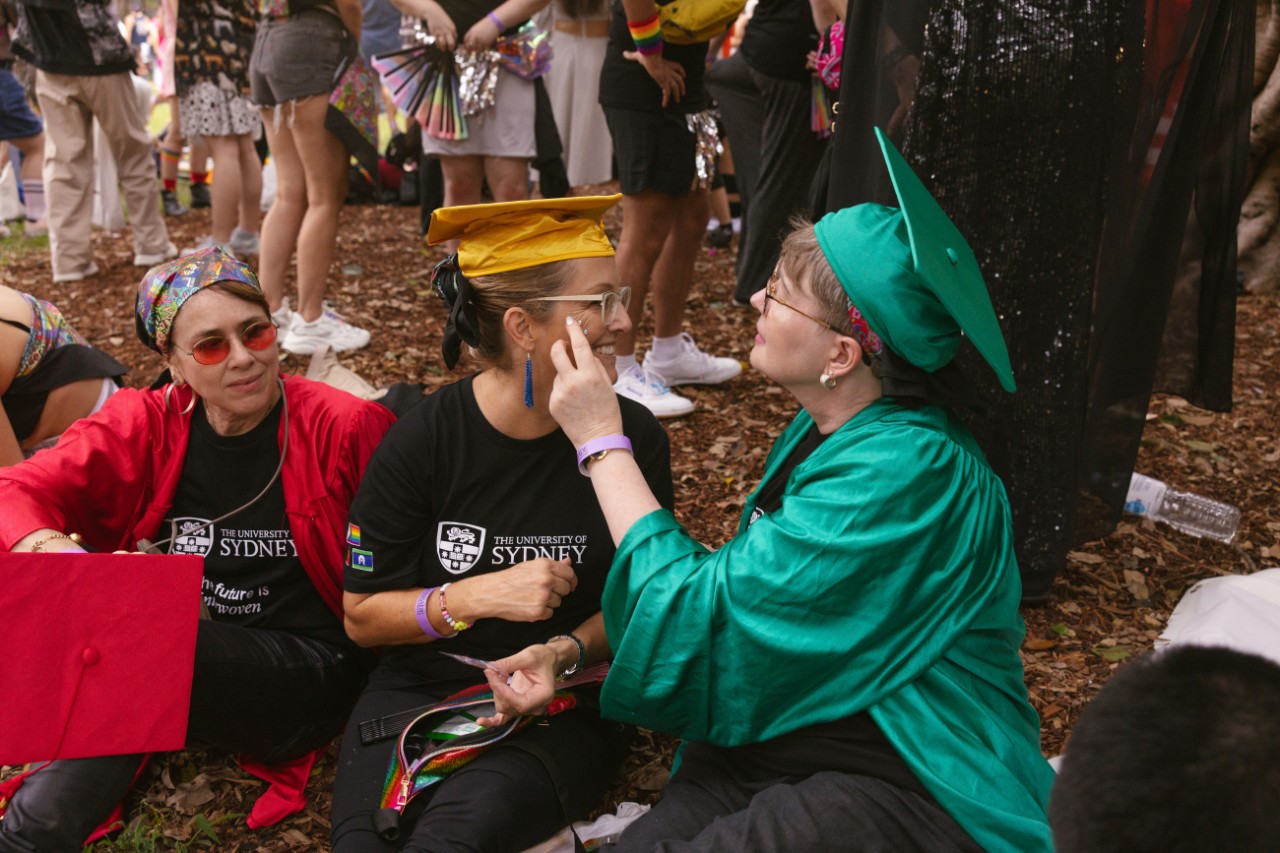 Three people sitting in a park. One is applying gemstone decorations to other's face. 