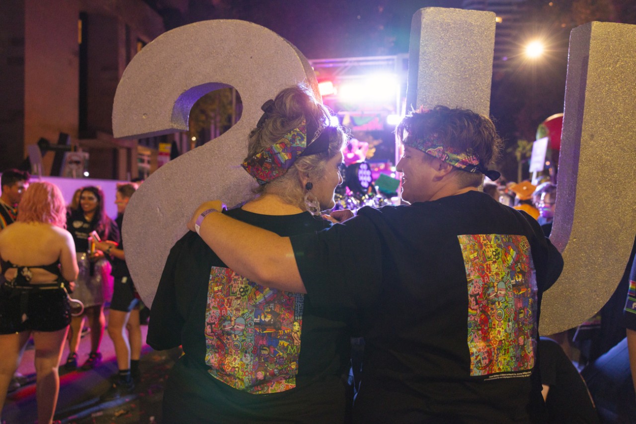 Two people with matching colourful t-shirts stand together, one with their arm around the other.