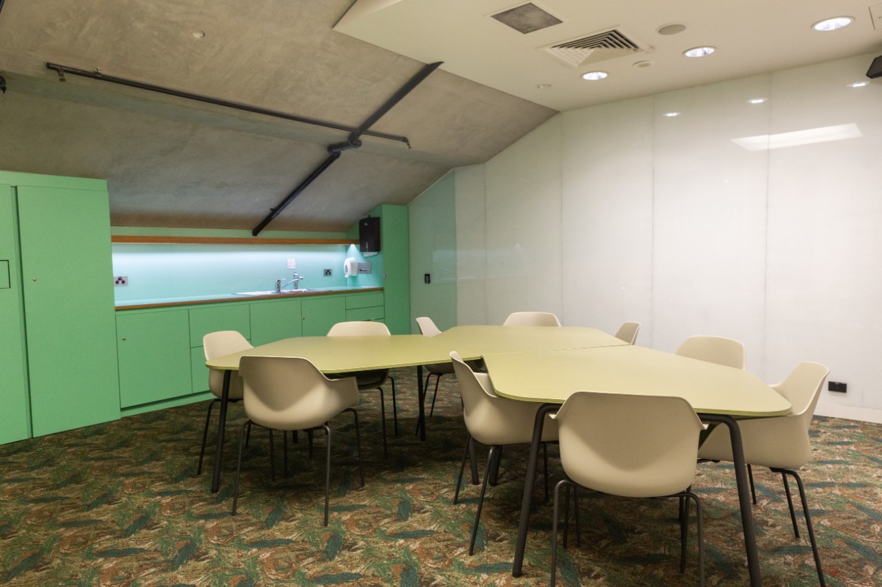 Room with desks surrounded by chairs, with a kitchenette along the side 