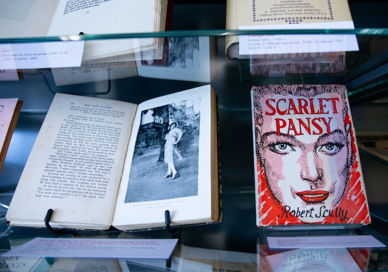 Two books side by side. One. Man Into Woman, is open to a photograph of Lili Elbe. The other, Scarlet Pansy, is closed, and features a striking red cover image of an androgynous face