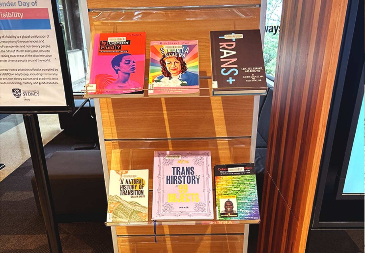 a book case displaying 6 books on trans themes, with a stand next to it promoting the display