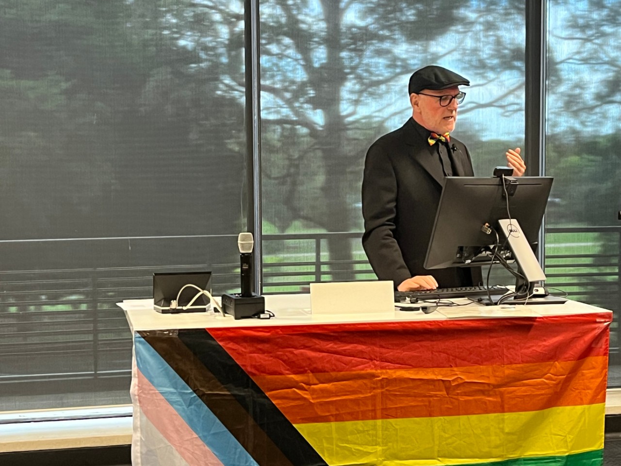 A colour photograph showing Vince Mitchell (a white man wearing a black suit and a rainbow bow tie) presenting at a lectern decorated with a progress pride flag.