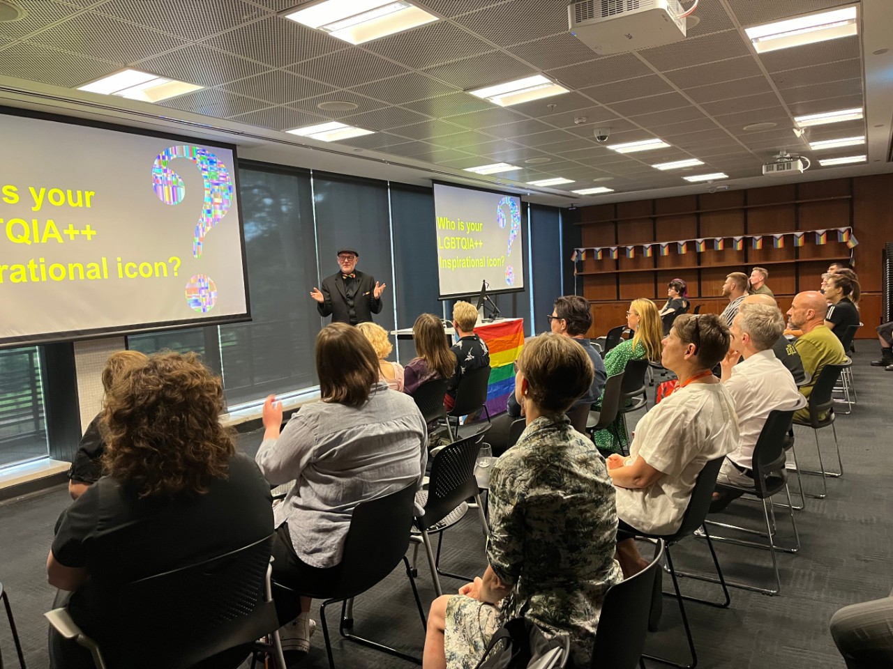 A colour photograph showing Professor Vince Mitchell (a white man wearing a black suit and rainbow bow tie) presenting to a room full of people. The projection screens behind him are displaying a slide with the words "Who is your LGBTQIA++ icon?", and the room is decorated with rainbow pride flags