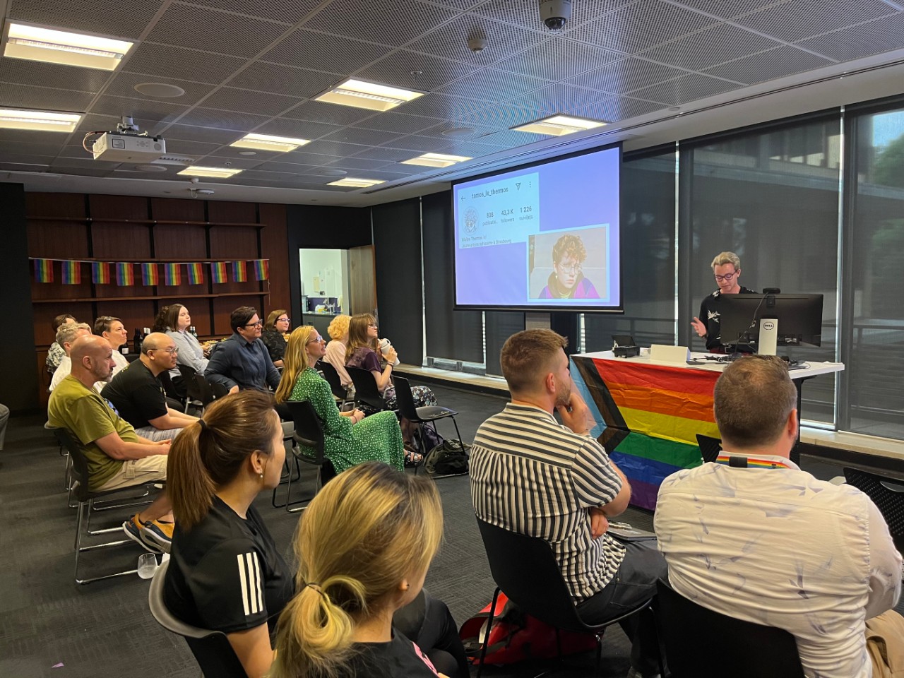 A photograph showing Jay Boyd (a young man with blonde hair and glasses) presenting to a room full of people. The screen behind him is showing the instagram profile of French graphic novelist Thamos le Thermos, and the room is decorated with rainbow pride flags.