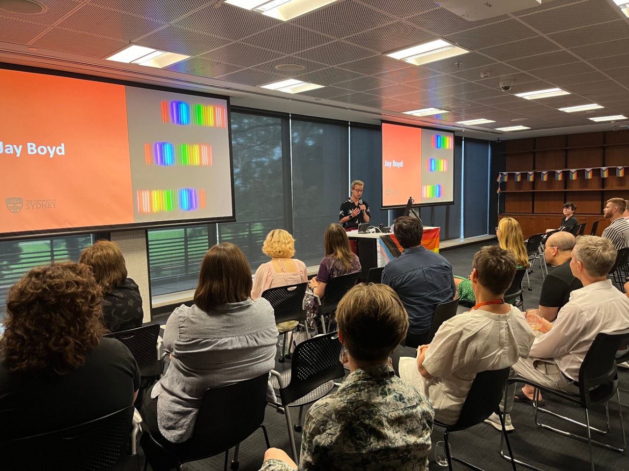 A photograph showing Jay Boyd (a young man with blond hair and glasses) presenting to a room full of people. The projection screens behind him are displaying his name, and the room is decorated with rainbow pride flags.