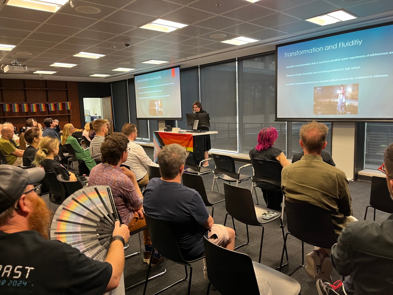 A photograph showing Lauren Mills (a fair skinned person with glasses) presenting to a room full of people. The projection screens behind them are displaying a slide reading "Transformation and Fluidity". In the foreground, someone is holding a rainbow fan.
