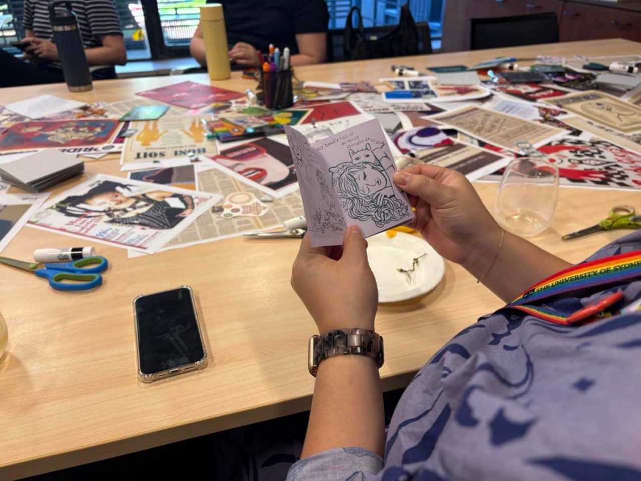 A photograph taken over someone's shoulders, showing them holding a small zine (DIY magazine) in their hands. On the table in front of them is a variety of craft materials