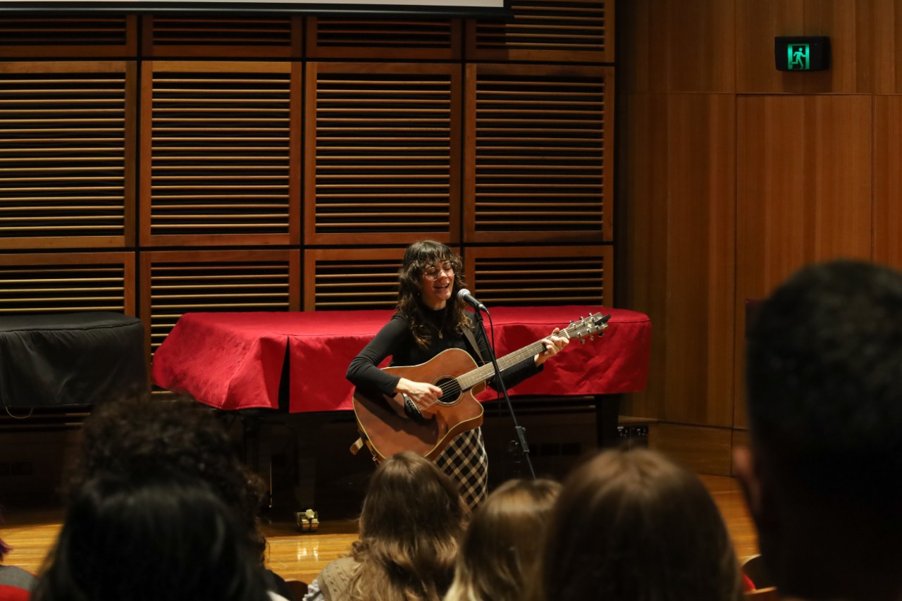 A colour photograph showing Caitlin Erbacher (a white woman with brunette hair and glasses) singing and playing acoustic guitar in front of an audience.