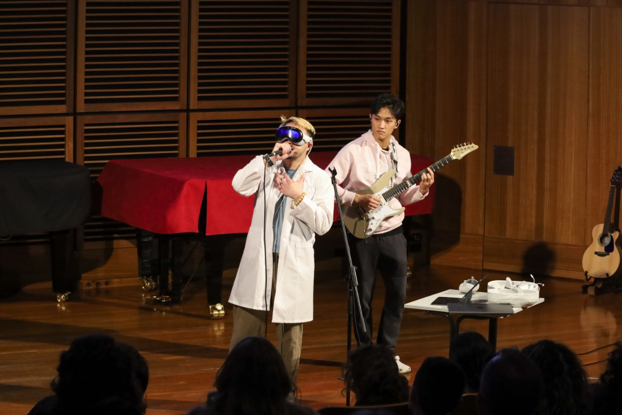 A colour photograph showing two Asian men performing in front of an audience. One of the men (Bohan) is singing and wearing virtual reality goggles and a lab coat. The other (Terry) is playing electric guitar and wearing a light pink hoodie.