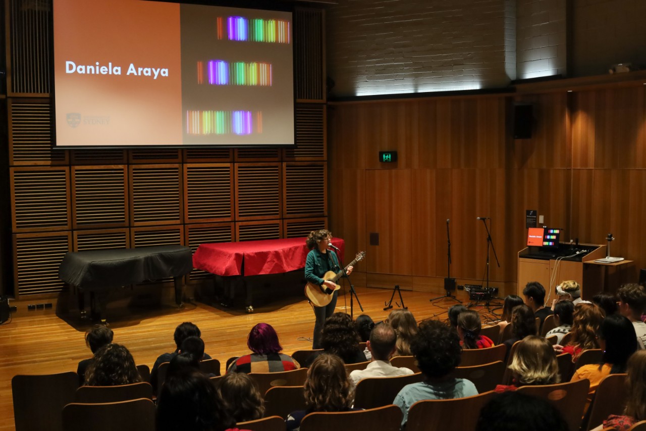 A colour photograph showing a Latina woman with brunette hair and glasses singing and playing acoustic guitar in front of a live audience. Behind her is a large display screen that reads "DANIELA ARAYA".