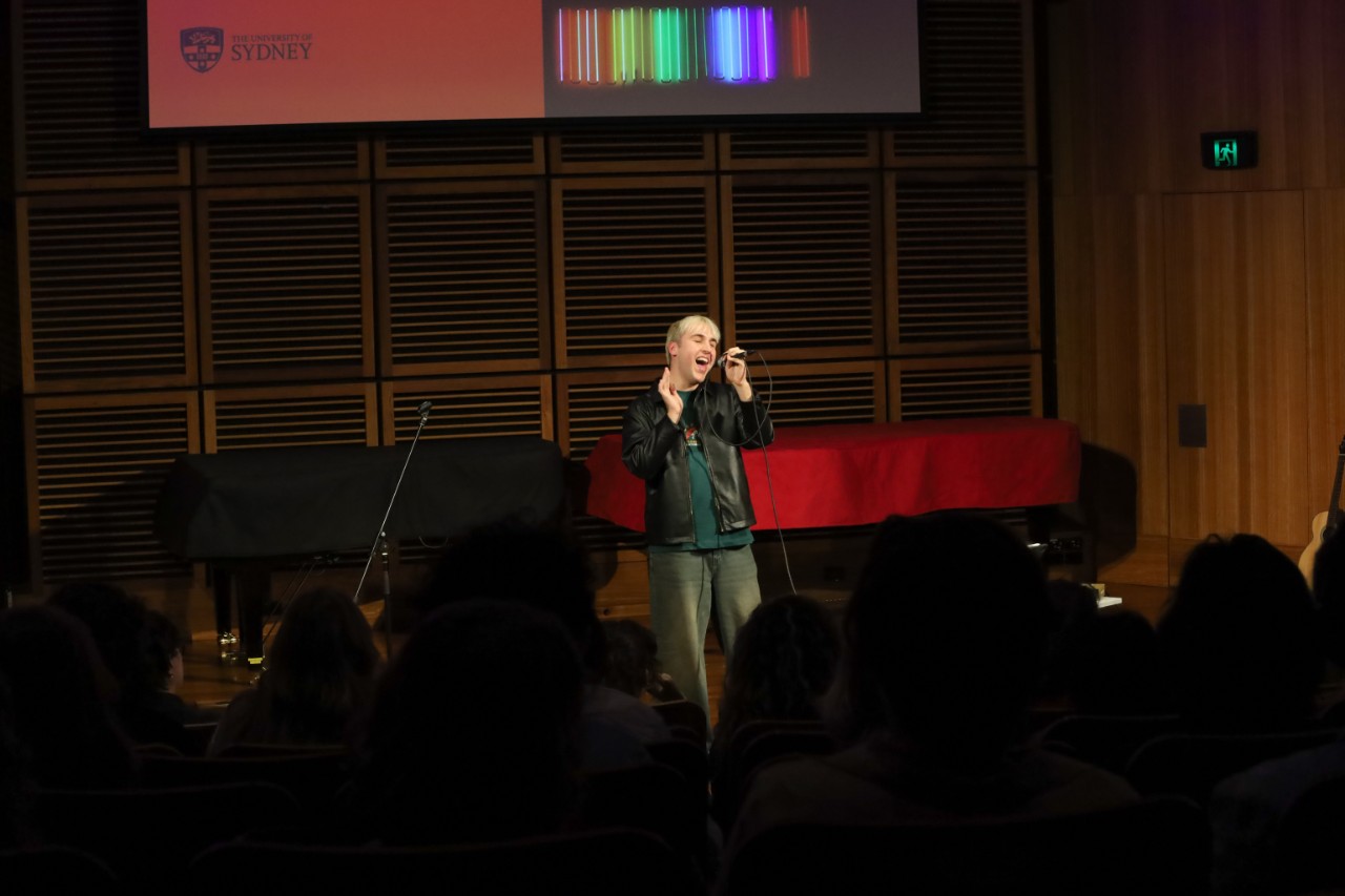 A colour photograph showing Aleks Matic (a white man with platinum blonde hair) singing into a microphone in front of an audience.