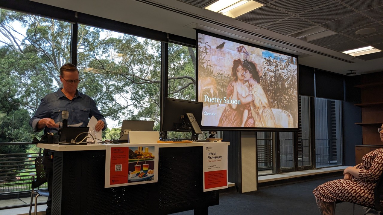 A photograph showing a fair-skinned person with short hair presenting at a lectern. In the background is a projector screen showing a presentation slide that reads "Poetry Saloon: open mic"