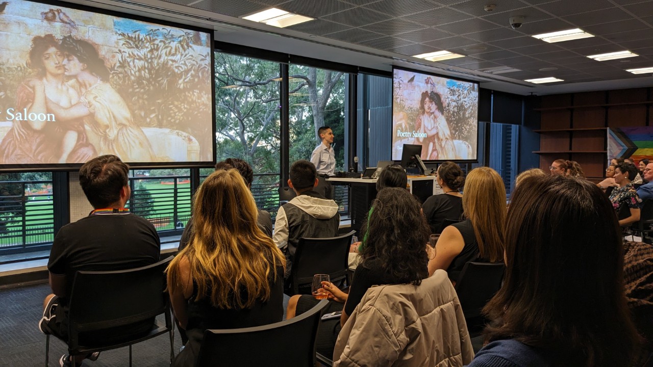 A photograph showing a fair-skinned person with short hair presenting at a lectern. In the foreground is an audience of several dozen people listening attentively to the speaker. In the background is a projector screen showing a presentation slide that reads "Poetry Saloon: open mic"
