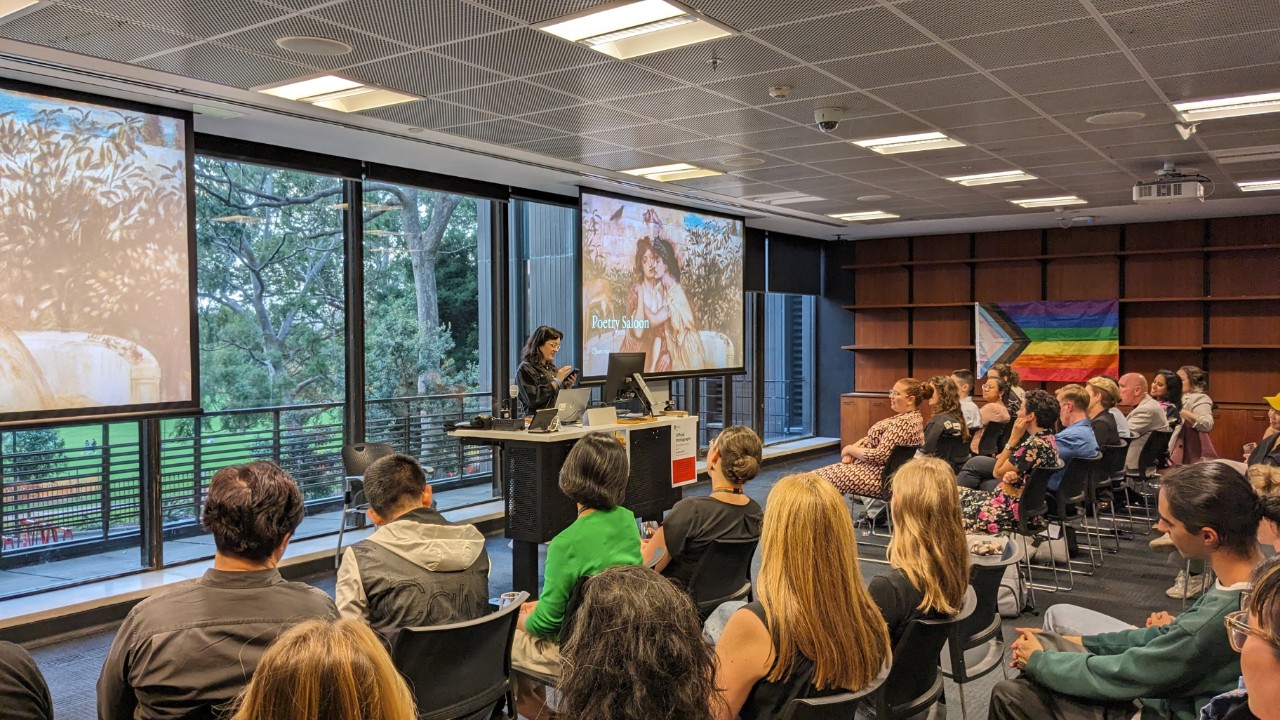A photograph showing a fair-skinned person with long brown hair and glasses presenting at a lectern to an attentive audience. In the background is an LGBTQIA+ pride progress flag and a projector screen showing a presentation slide that reads "Poetry Saloon: open mic"