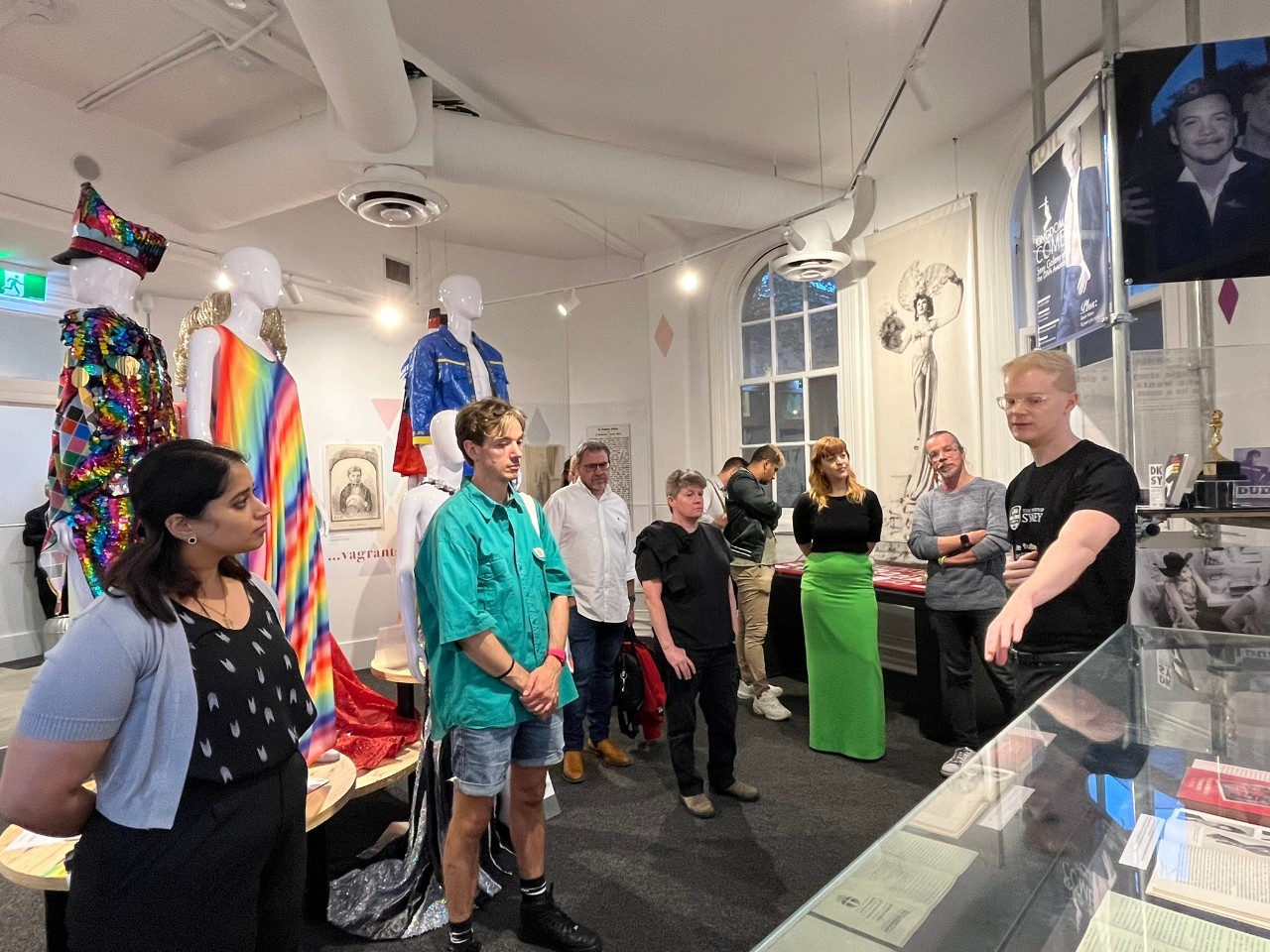 A photograph showing a group of people in a brightly lit exhibition space. On the right hand side of the frame, a white man with blonde hair and glasses is speaking and gesturing towards one of the display cases while the rest of the people in the room look and listen. In the backgroud, several mannequins wearing brightly coloured and sequinned drag costumes can be seen.