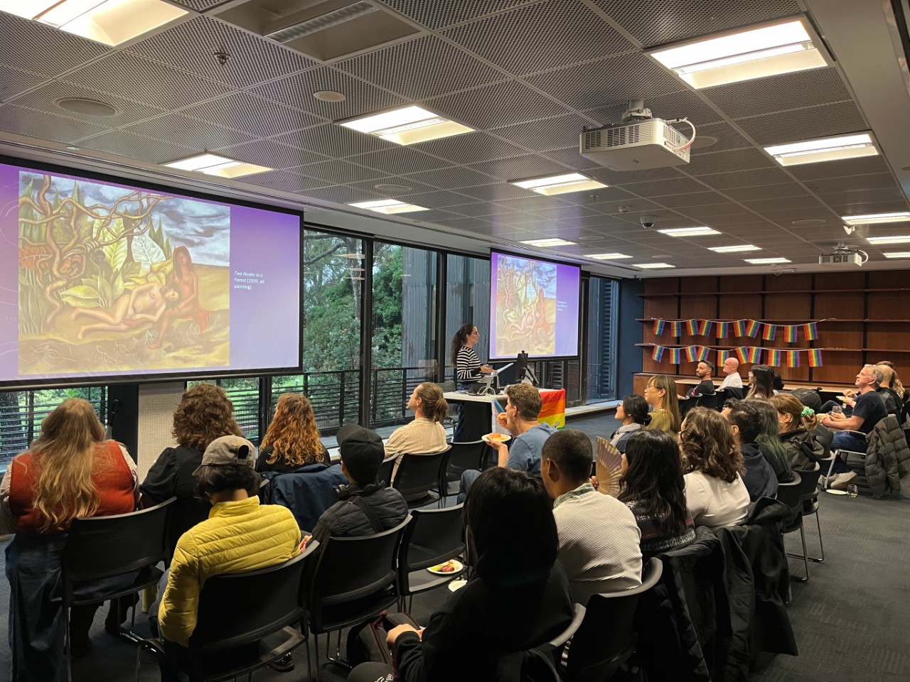 A colour photograph showing Alexandra Angus (a white woman with glasses and long brunette hair in a ponytail) presenting to a room full of people. The projection screens behind her are displaying Frida Kahlo's "Two Nudes in a Forest" and the room is decorated with rainbow pride flags