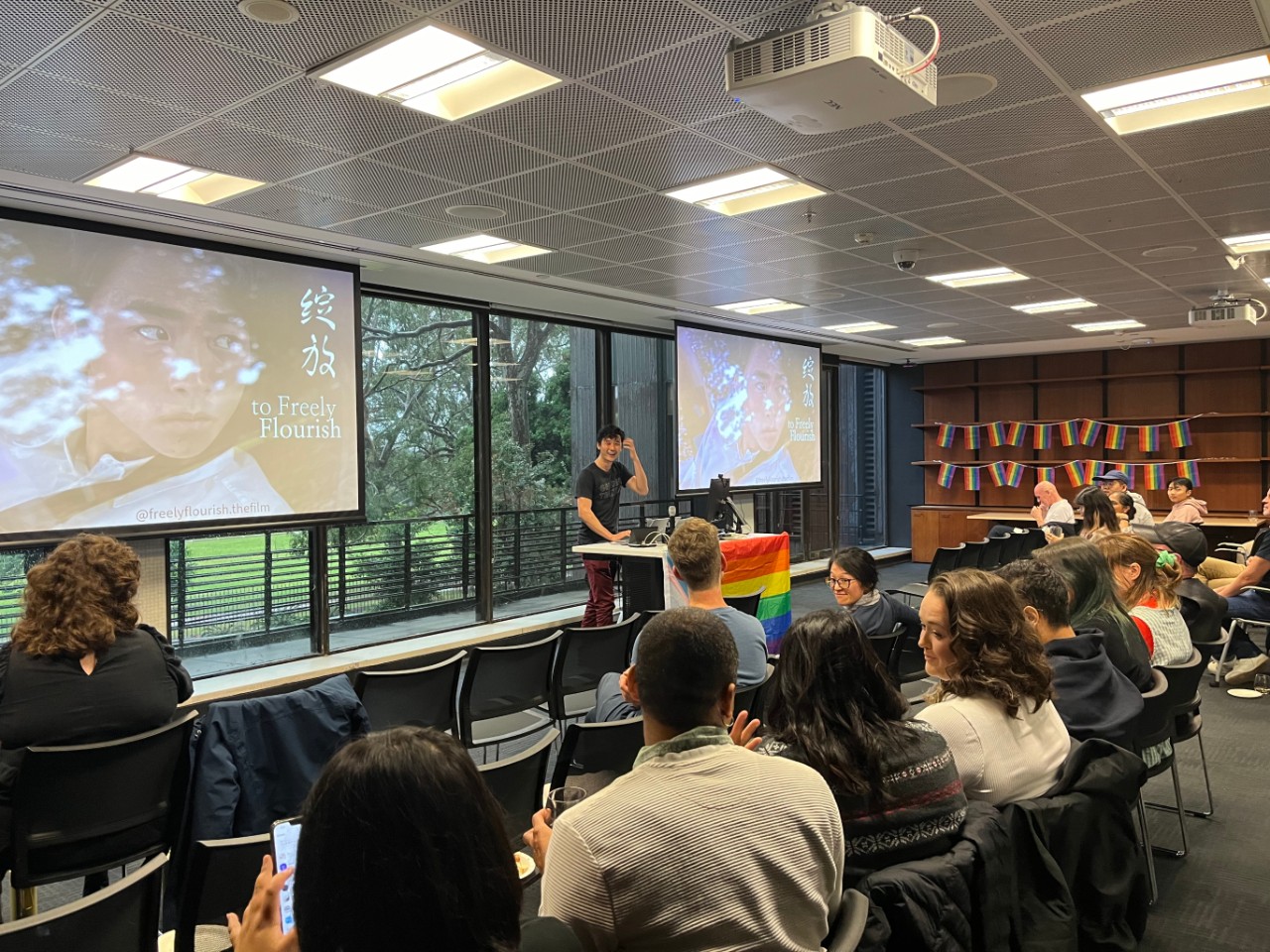 A colour photograph showing Victor Wu, a Chinese Australian man, smiling and presenting to a room full of people. The projection screens behind him are displaying a still from his film To Freely Flourish, and the room is decorated with rainbow pride flags