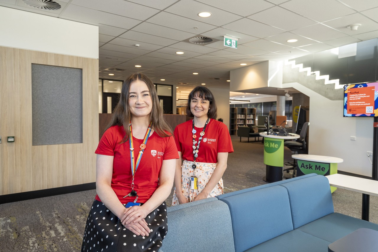 Two library staff in red T-shirts stand inside the entrance of the Susan Wakil Health Building Library, with the information desk and book shelves visible in the background